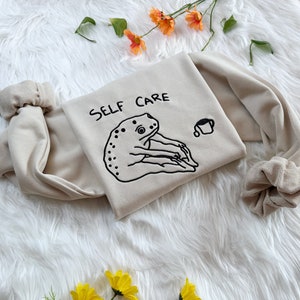 May include: A beige sweatshirt with the text "SELF CARE" embroidered in black. A black and white illustration of a frog holding a cup is embroidered below the text.