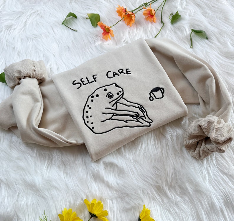 May include: A beige sweatshirt with the text "SELF CARE" embroidered in black. A black and white illustration of a frog holding a cup is embroidered below the text.