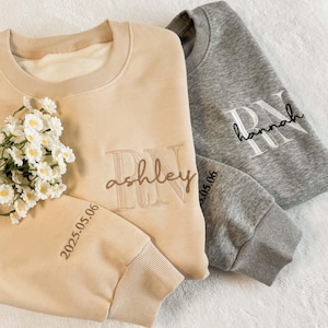 May include: Two crewneck sweatshirts, one beige and one grey, with embroidered details. The beige sweatshirt features "RN ashley" and the date "2025.05.06" in brown thread. The grey sweatshirt has "RN hannah" in white thread and the date "05.06".