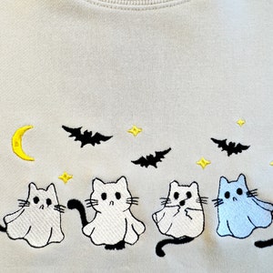 May include: A light beige sweatshirt with an embroidered design of four cats dressed as ghosts. The cats are white with black eyes and tails, and one cat is light blue. The design also includes a crescent moon, stars, and bats.