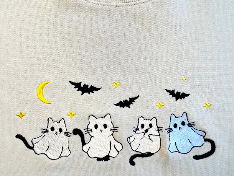 May include: A light beige sweatshirt with an embroidered design of four cats dressed as ghosts. The cats are white with black eyes and tails, and one cat is light blue. The design also includes a crescent moon, stars, and bats.