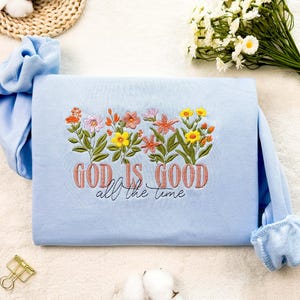 Könnte beinhalten: Hellblauer Pullover mit gestickten Blumen und dem Text "God is good all the time".