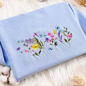 Può includere: Una felpa blu chiaro con un ricamo di fiori e farfalle colorati.