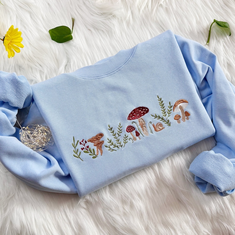 Op de afbeelding: Een lichtblauw sweatshirt met een geborduurd ontwerp van paddenstoelen en bladeren. Het ontwerp is in tinten bruin, rood en groen.