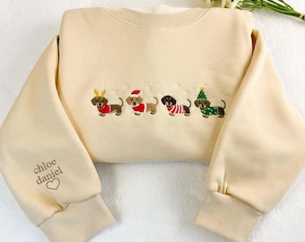 Puede incluir: Sudadera color crema con bordados de perros salchicha con atuendos festivos. La manga izquierda tiene el texto "chloe daniel" con un corazón. La sudadera tiene cuello redondo y puños acanalados.