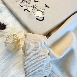 May include: A beige sweatshirt with an embroidered design of four ghost cats and bats. The sweatshirt has a ribbed cuff and is folded in half.