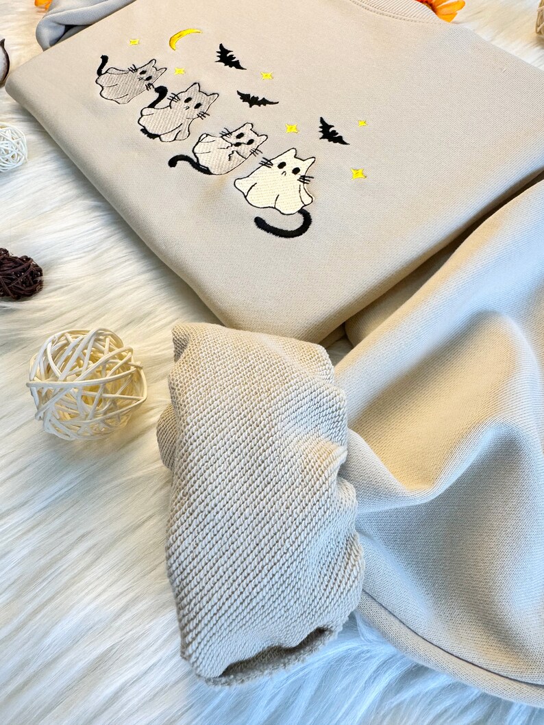May include: A beige sweatshirt with an embroidered design of four ghost cats and bats. The sweatshirt has a ribbed cuff and is folded in half.