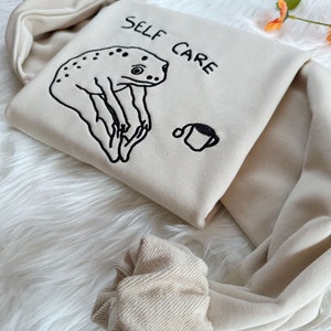May include: A beige hoodie with a frog illustration and the text "SELF CARE" embroidered on the front. The hoodie has a drawstring hood and ribbed cuffs.