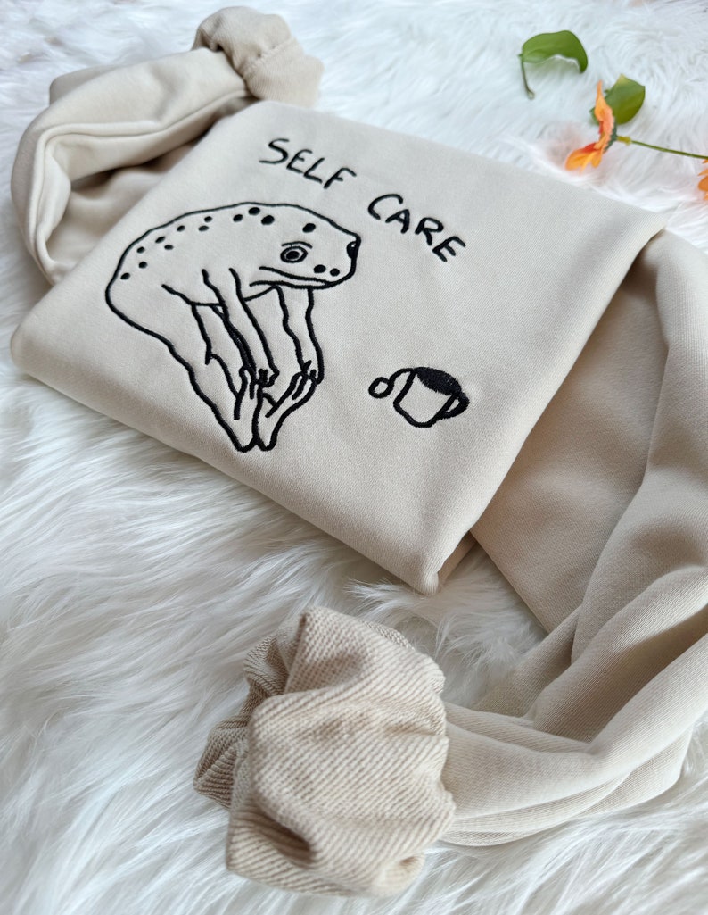 May include: A beige hoodie with a frog illustration and the text "SELF CARE" embroidered on the front. The hoodie has a drawstring hood and ribbed cuffs.