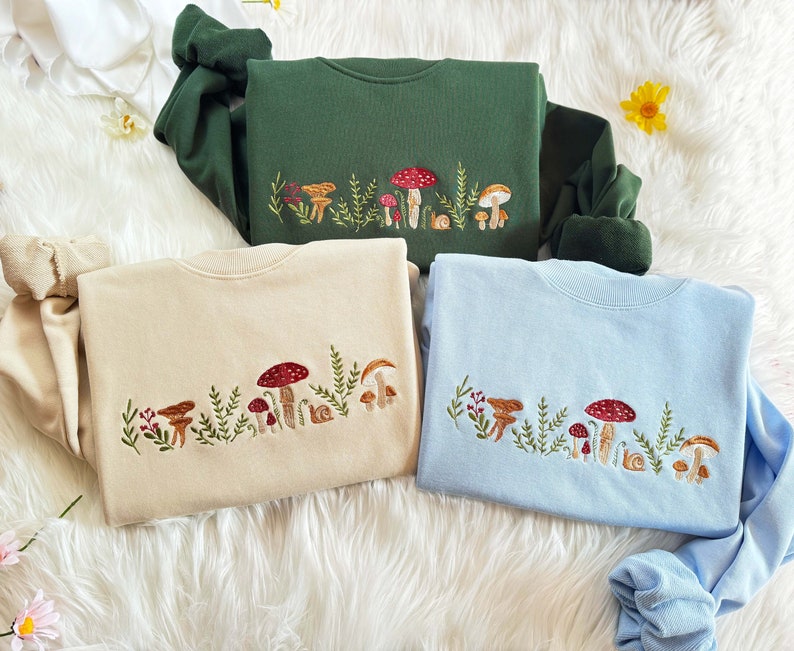Op de afbeelding: Drie sweatshirts met geborduurde paddenstoelontwerpen. De sweatshirts zijn in groen, beige en lichtblauw.