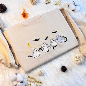 May include: A beige sweatshirt with an embroidered design of four white ghost cats with yellow eyes and black noses. The cats are arranged in a line, with the largest cat at the back and the smallest cat at the front. The design also includes a crescent moon, stars, and bats.