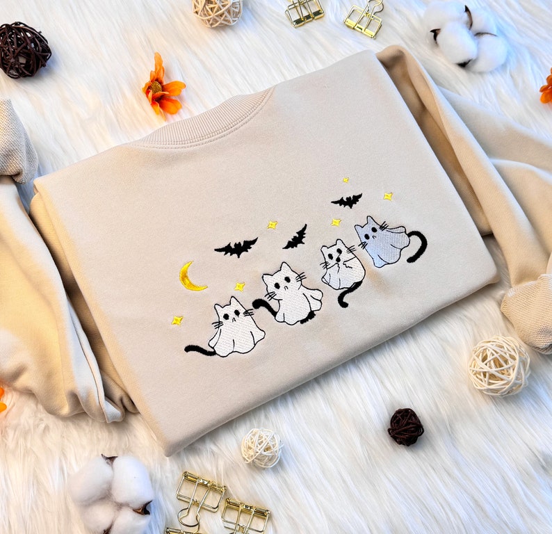 May include: A beige sweatshirt with an embroidered design of four white ghost cats with yellow eyes and black noses. The cats are arranged in a line, with the largest cat at the back and the smallest cat at the front. The design also includes a crescent moon, stars, and bats.