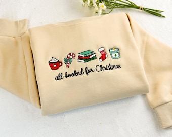 Pode incluir: Camisola bege com ícones bordados com tema natalino, incluindo uma caneca, bengala doce, livros, meia e vela. A frase "all booked for Christmas" está bordada abaixo. A camisola tem mangas compridas.