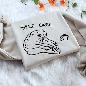May include: A beige sweatshirt with the text "SELF CARE" embroidered in black. A black and white illustration of a frog is embroidered below the text. A small black and white illustration of a cup of coffee is embroidered on the right side of the sweatshirt.