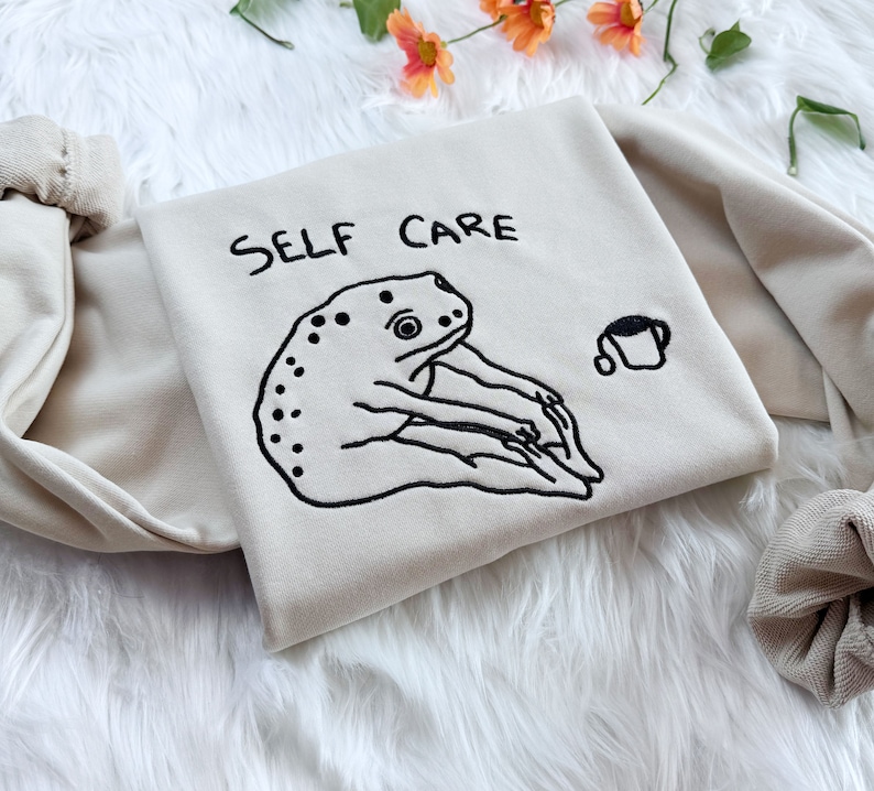May include: A beige sweatshirt with the text "SELF CARE" embroidered in black. A black and white illustration of a frog is embroidered below the text. A small black and white illustration of a cup of coffee is embroidered on the right side of the sweatshirt.