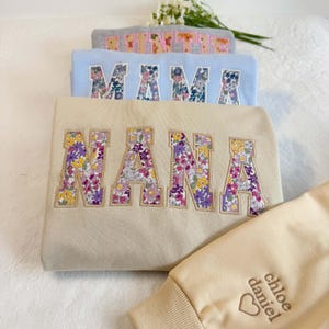 May include: Several sweatshirts in a stack, each embroidered with a floral word. The top one is grey with "MAMA", followed by a light blue one with "MAMA", and a beige one with "NANA". A beige sleeve is visible with the names "chloe daniel" and a heart.