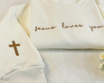 May include: Cream-colored sweatshirt with the embroidered phrase "jesus loves you" in brown script. A brown cross is embroidered on the left sleeve. The sweatshirt has a ribbed collar and cuffs.