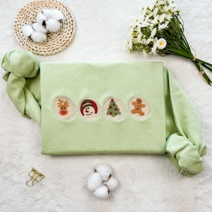 May include: A light green sweatshirt with four embroidered Christmas-themed designs: a reindeer, a snowman, a Christmas tree, and a gingerbread man.