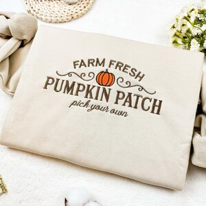 Puede incluir: Sudadera beige con la frase bordada "FARM FRESH PUMPKIN PATCH pick your own" y un gráfico de calabaza naranja. La prenda está doblada sobre una superficie blanca.