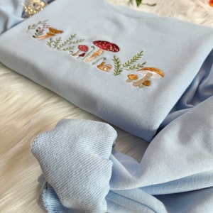 Op de afbeelding: Een lichtblauw sweatshirt met een geborduurd ontwerp van paddenstoelen en bladeren. Het ontwerp is in tinten bruin, rood en wit.