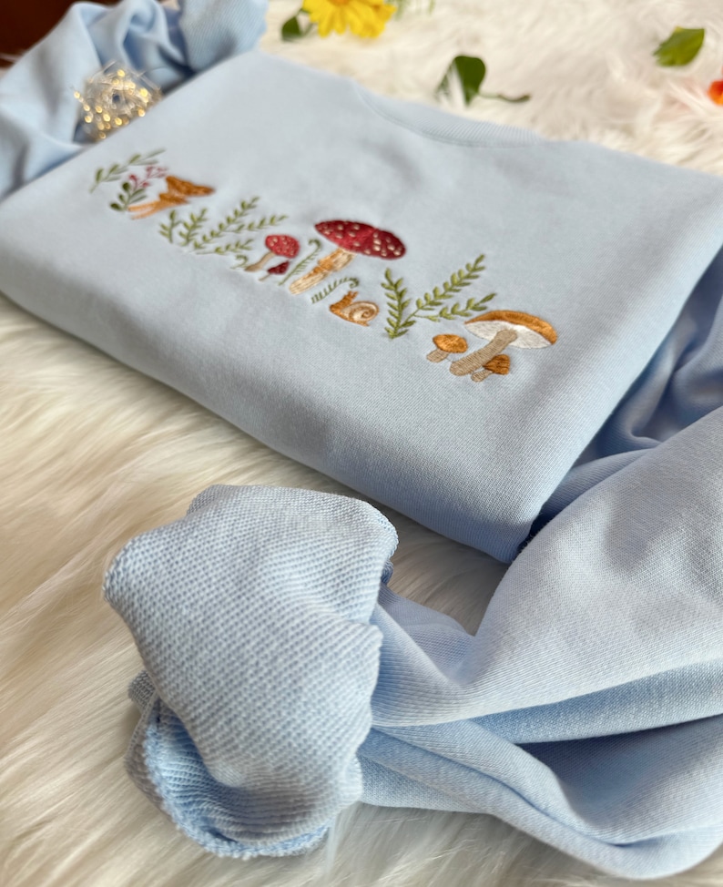 Op de afbeelding: Een lichtblauw sweatshirt met een geborduurd ontwerp van paddenstoelen en bladeren. Het ontwerp is in tinten bruin, rood en wit.