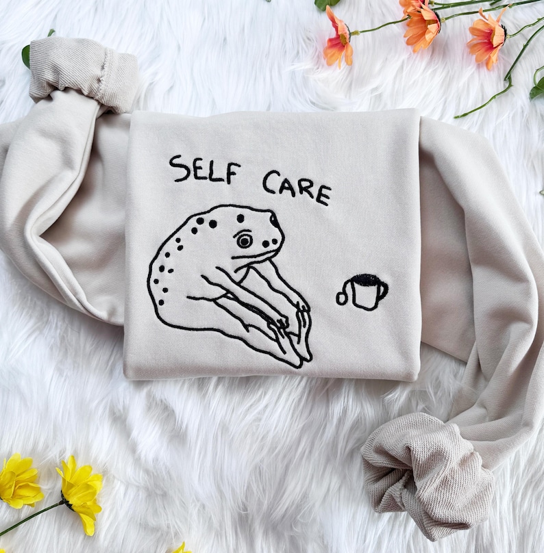 May include: A beige sweatshirt with the text "SELF CARE" embroidered in black. A black and white illustration of a frog doing yoga is embroidered below the text. A small black and white illustration of a cup is embroidered to the right of the frog.
