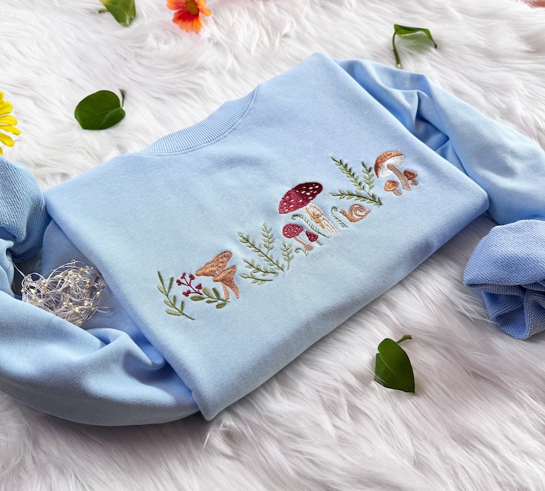 Op de afbeelding: Een lichtblauw sweatshirt met een geborduurd ontwerp van paddenstoelen, bladeren en een slak. Het ontwerp is in tinten bruin, rood en groen.
