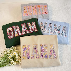 May include: Five sweatshirts in different colours, each embroidered with a name in floral print lettering. The names include "AUNTIE", "GRAM", "MAMA", and "NANA". The sweatshirts are in shades of grey, green, blue, and beige.