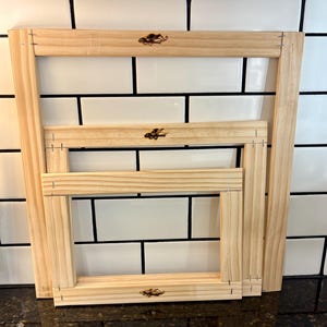 May include: Three unfinished wooden frames of different sizes are stacked. The frames are rectangular with mitred corners, held together with staples. Each frame has a small, dark logo in the upper right corner.