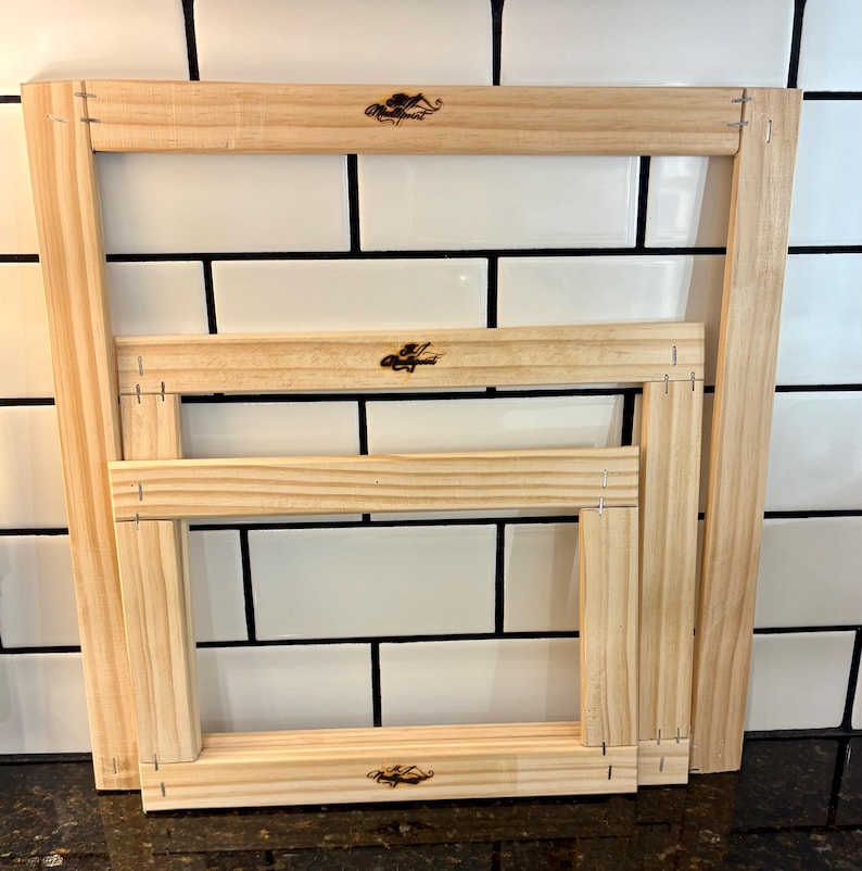 May include: Three unfinished wooden frames of different sizes are stacked. The frames are rectangular with mitred corners, held together with staples. Each frame has a small, dark logo in the upper right corner.