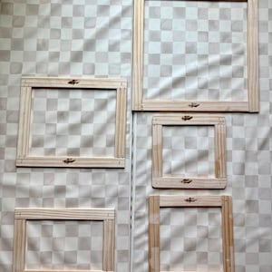 May include: Five wooden embroidery hoops of different sizes are displayed against a patterned backdrop. Each hoop is rectangular and crafted from light-coloured wood, featuring decorative metal clasps. The hoops are empty, ready for fabric to be stretched across.