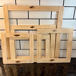 May include: Four unfinished wooden embroidery hoops of different sizes. Each rectangular hoop has a decorative emblem and is constructed with visible staples. The hoops are displayed against a white tiled background.
