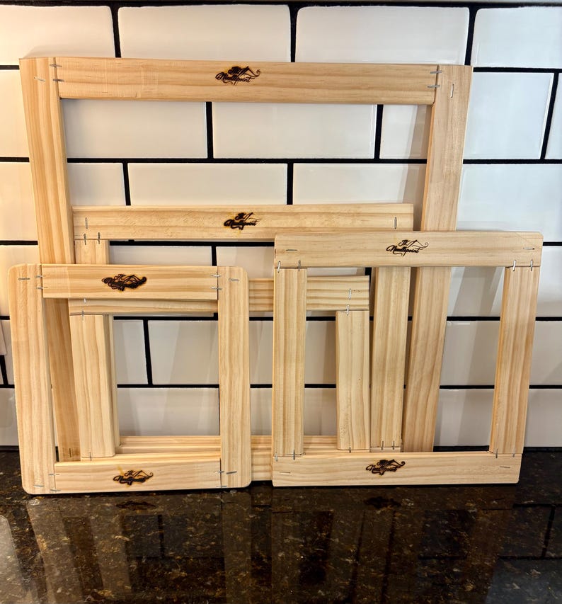 May include: Four unfinished wooden embroidery hoops of different sizes. Each rectangular hoop has a decorative emblem and is constructed with visible staples. The hoops are displayed against a white tiled background.
