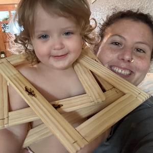 May include: A smiling toddler holds several unfinished wooden canvas frames. The frames are made of light-coloured wood and are interlocked. The child is wearing a nappy. An adult is smiling behind the child.