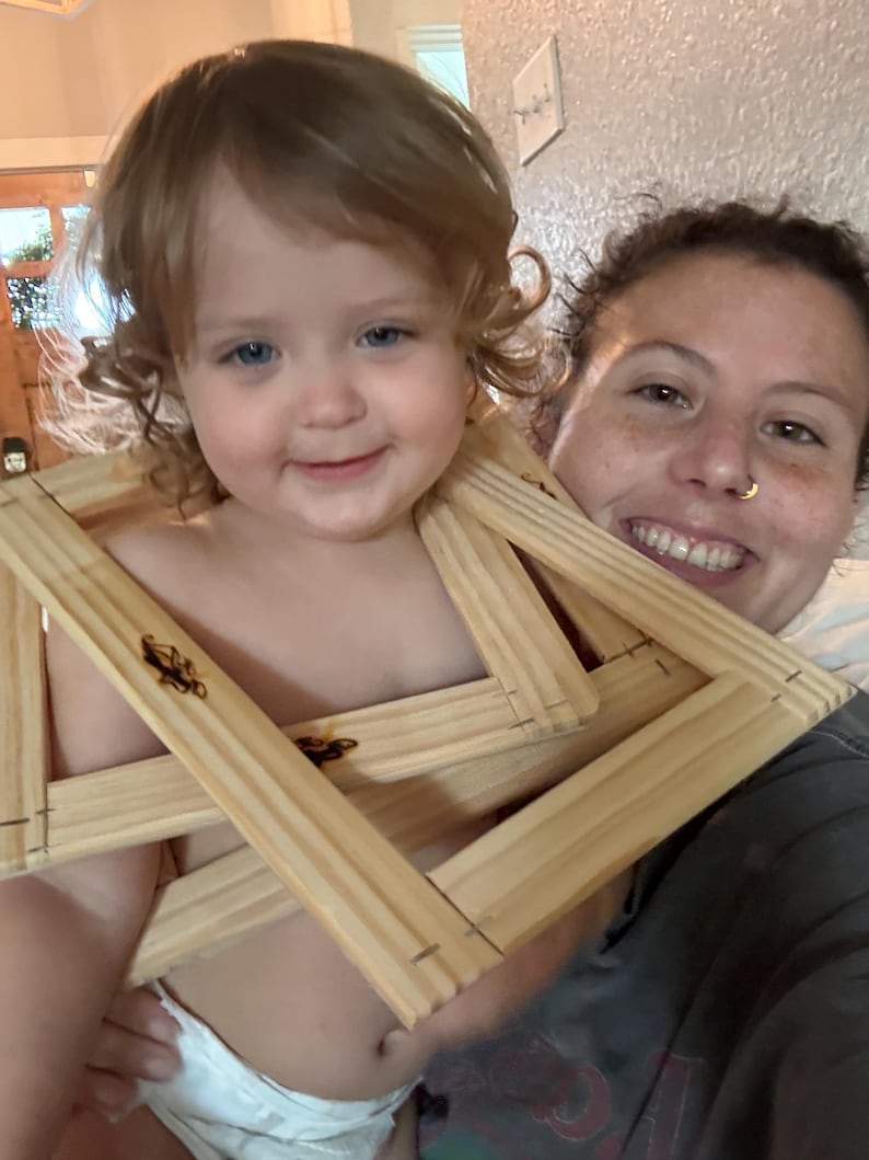 May include: A smiling toddler holds several unfinished wooden canvas frames. The frames are made of light-coloured wood and are interlocked. The child is wearing a nappy. An adult is smiling behind the child.