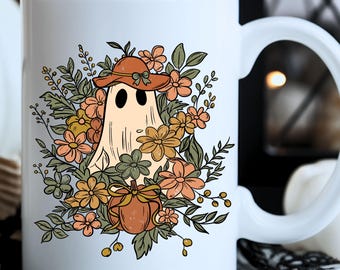 May include: White ceramic mug featuring a whimsical ghost design. The ghost wears a hat and is surrounded by flowers in shades of orange, peach, and green. A pumpkin is also included in the design. The mug has a curved handle.