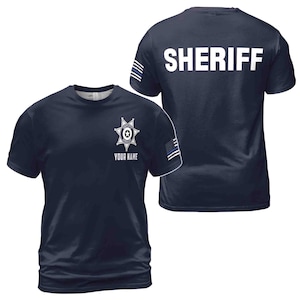 May include: A navy blue t-shirt with a white sheriff's star badge on the front and the word "SHERIFF" printed on the back. The shirt has a thin blue line on the sleeve.