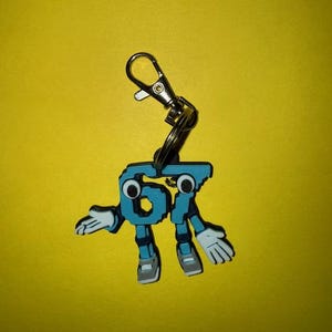 May include: A blue and white keychain in the shape of the number 67 with cartoon-like arms, legs, and eyes. The keychain has a silver clasp and ring. The background is a solid yellow.