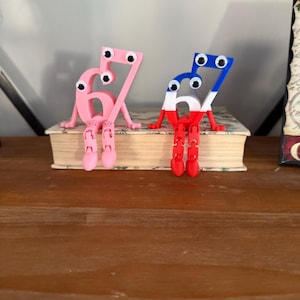 May include: Two whimsical, 3D-printed figures of the numbers 6 and 7 with googly eyes, arms, and legs. One is pink, the other is red, white, and blue. They are sitting on a book, adding a playful touch to any space.