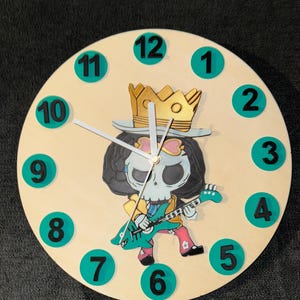 May include: A round wooden clock with a cartoon character design. The clock face is light beige with teal numbers and hands. The central image is a character with a skull face, wearing a crown and playing a teal guitar.