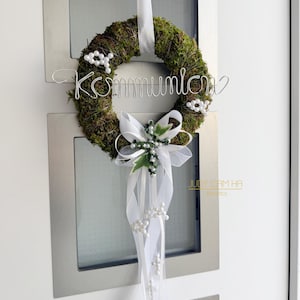 Könnte beinhalten: Ein runder Mooskranz mit dem Wort "Kommunion" aus silbernem Draht. Er ist mit weißen Blumen, einer weißen Schleife und einem langen weißen Band mit Perlen verziert. Der Kranz hängt an einer Tür.