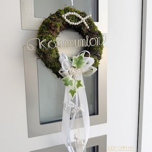 Könnte beinhalten: Ein mit Moos bedeckter Kranz mit dem Wort "Kommunion" aus silbernem Draht. Der Kranz ist mit einem Fischsymbol aus Perlen, weißen Bändern, künstlichem Efeu und einer herzförmigen Verzierung geschmückt. Ein dekoratives Stück für einen besonderen Anlass.
