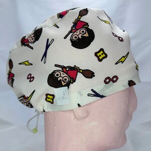 May include: White surgical scrub hat with a repeating pattern of Harry Potter characters, lightning bolts, glasses, and wands. The hat has a drawstring and a small label that says "Blue Flowers".