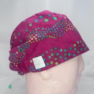 May include: Pink surgical scrub hat with a colourful, abstract pattern of dots and swirls. The hat has a drawstring and a small white label with the text "Happy Flowers".