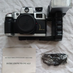 Può includere: Una macchina fotografica a pellicola da 35 mm argento e nero con un obiettivo nero e un flash incorporato. La macchina fotografica ha una funzione di autoscatto e viene fornita con una cinghia nera e istruzioni in italiano.
