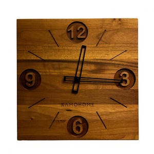 Puede incluir: Un reloj de pared de madera con manecillas negras y números romanos. La esfera del reloj está hecha de madera con un patrón de veta natural. El reloj está etiquetado como "KAMO HOME".