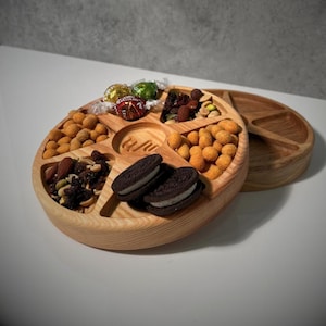 Könnte beinhalten: Ein runder Servierteller aus hellem Holz mit mehreren Fächern, gefüllt mit verschiedenen Snacks. Das Tablett enthält Erdnüsse, Kekse, Nüsse und verpackte Süßigkeiten. Ein zweites, ähnliches Tablett ist im Hintergrund teilweise sichtbar.