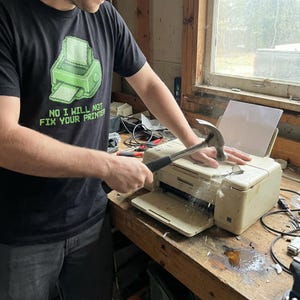 Könnte beinhalten: Eine Person zerstört einen beigen Drucker mit einem Hammer auf einer hölzernen Werkbank. Die Person trägt ein schwarzes T-Shirt mit einer grünen pixeligen Druckergrafik und dem Text "NO I WILL NOT FIX YOUR PRINTER".