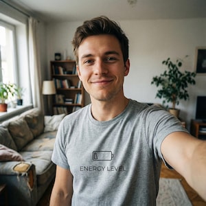 Könnte beinhalten: Ein graues T-Shirt mit dem Text "ENERGY LEVEL" unter einem Batteriesymbol. Die Person, die das T-Shirt trägt, lächelt und macht ein Selfie. Der Hintergrund umfasst ein Sofa, Bücherregale und ein Fenster.