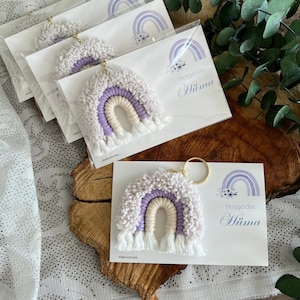 May include: A collection of handmade rainbow keychains in shades of lavender, cream, and white. Each keychain is attached to a gold ring and displayed on a card with a matching rainbow design and text.